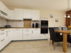 A modern kitchen with white cabinets, a built-in oven, and a dining area with a wooden table and black chairs. - Holiday Home in Grossenbrode (Grossenbrode)