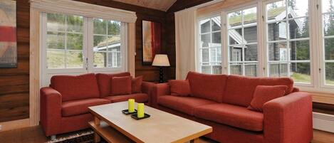 A cozy living room with a wooden ceiling, large windows, a red sofa set, a wooden coffee table, and a patterned rug.