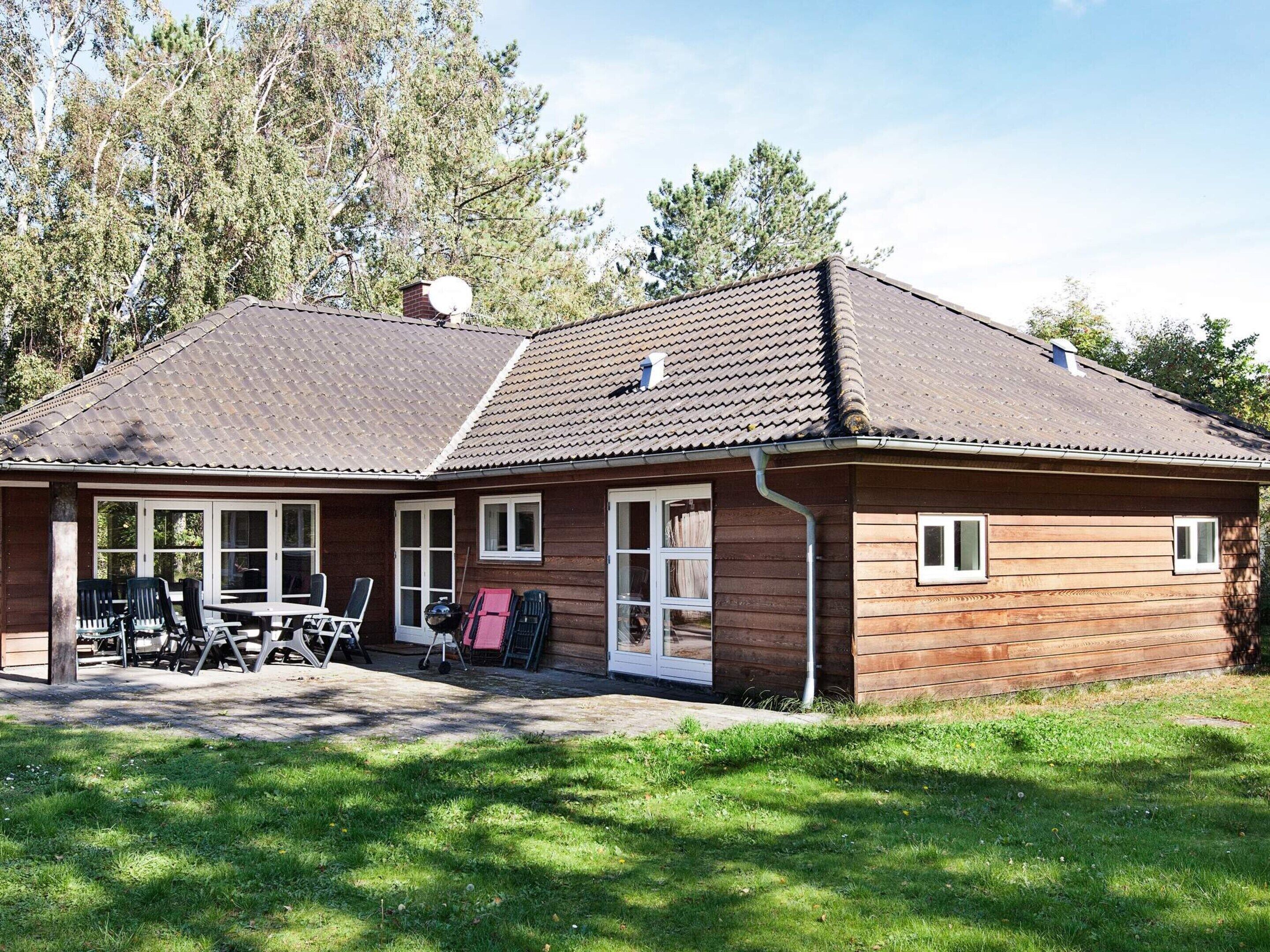 A wooden cabin with a tiled roof, a covered patio with chairs, and a grassy yard.