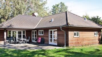 A wooden cabin with a tiled roof, a covered patio with chairs, and a grassy yard.