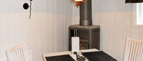 A room with a white table, two chairs, a wall-mounted lamp, and a wood-burning stove.
