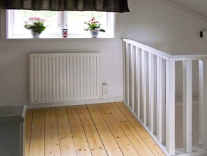 Une pièce avec un plancher en bois, des murs blancs, une fenêtre ornée de deux plantes en pot et une rambarde blanche.