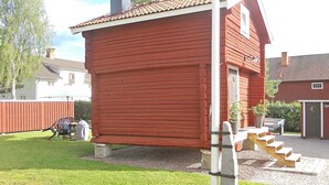 A red wooden building with a chimney, a staircase leading to a door, and a small garden area with a bench and a table. - 4 Person Holiday Home in Rattvik (Rattvik)