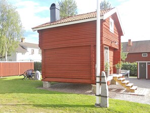 Un bâtiment en bois rouge avec une cheminée, un escalier menant à une porte et un petit jardin agrémenté d’un banc et d’une table.