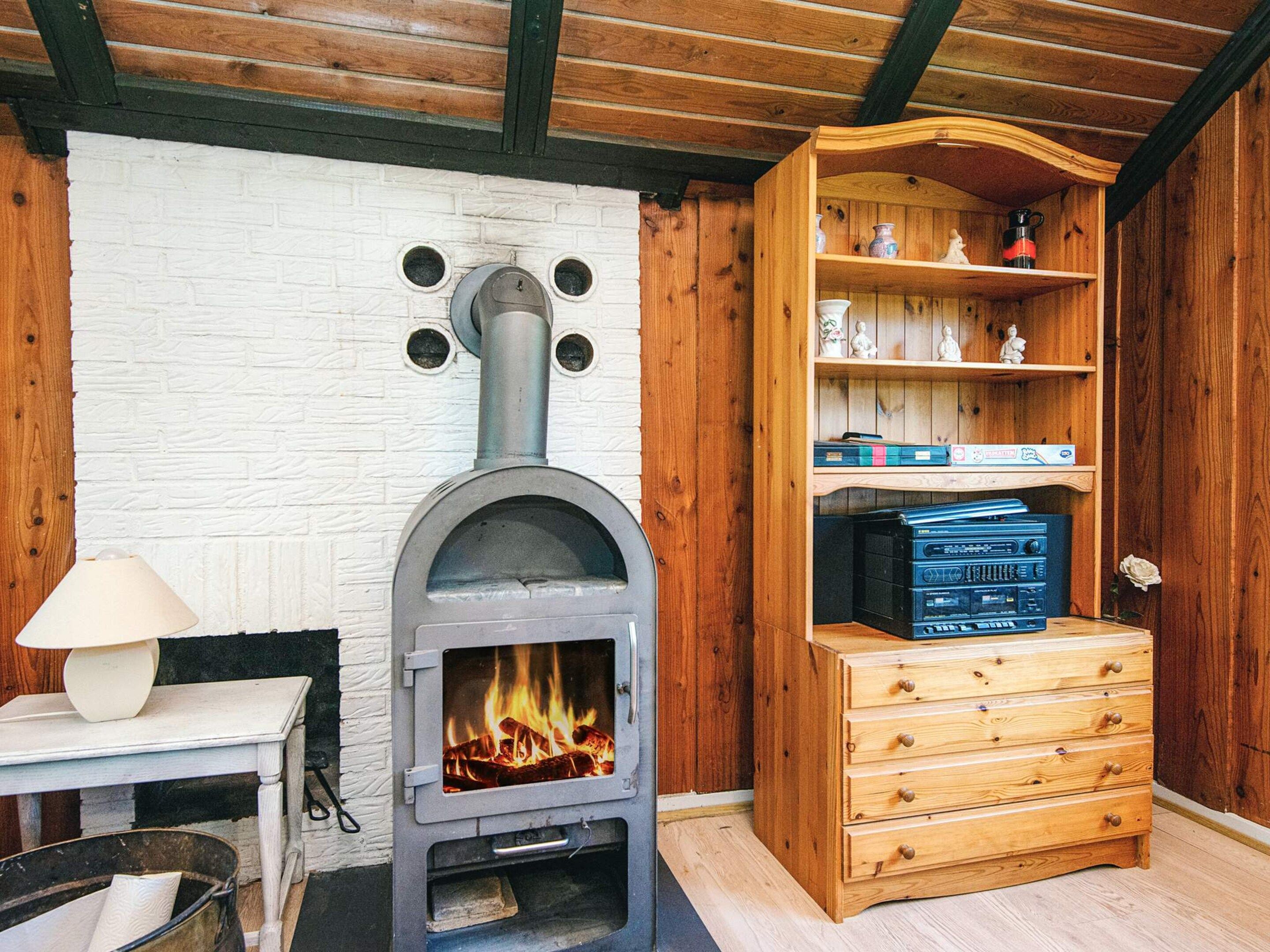A room with a wood-burning stove, a wooden cabinet with shelves, and a table with a lamp.