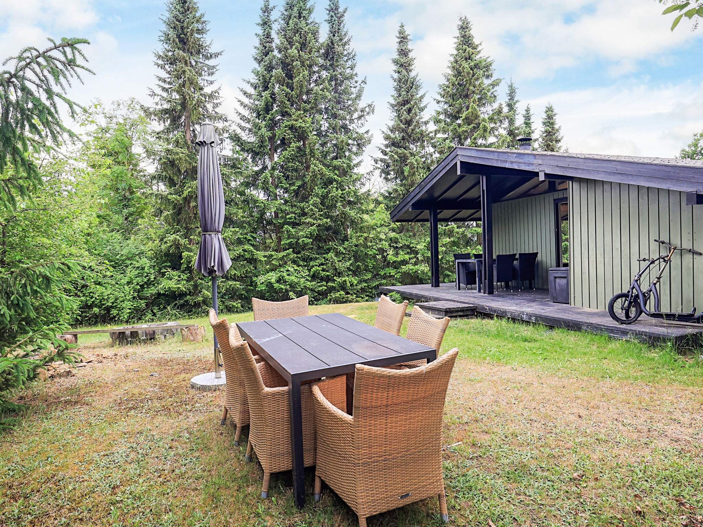 Une terrasse extérieure avec une table et des chaises, entourée de verdure et bordée d’un bâtiment doté d’une zone abritée.