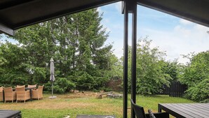 A covered outdoor deck with a table and chairs, surrounded by a grassy yard and trees.