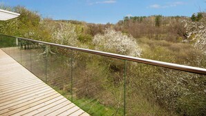 A wooden deck with a glass railing overlooking a natural landscape with trees and shrubs. - 8 Person Holiday Home in Ebeltoft-by Traum (Ebeltoft)
