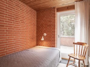 A room with a brick wall, a single bed with a quilted mattress, a wooden chair, a white lamp, and a sliding glass door leading to an outdoor area.