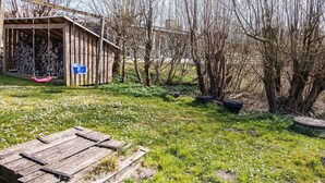 Un abri de jardin rustique en bois, agrémenté d’une boîte aux lettres bleue, est entouré d’une zone herbeuse parsemée de fleurs blanches et bordée d’arbres sans feuilles.