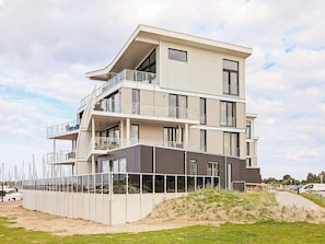 A multi-story residential building with balconies, large windows, and a sandy ground area.