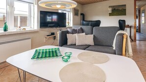Un salon moderne avec un canapé gris, un téléviseur à écran plat, un plafond en bois et une table blanche agrémentée d’objets décoratifs.