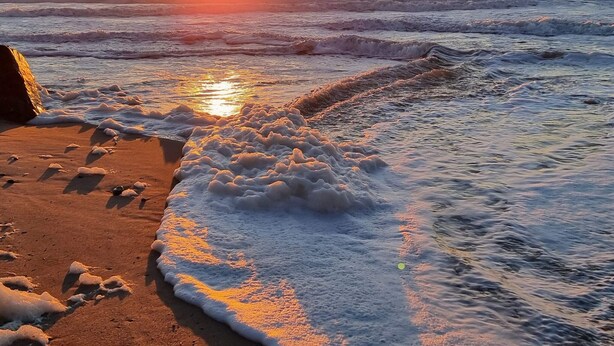 Une plage avec des vagues, un rocher et le soleil se couchant à l’horizon.