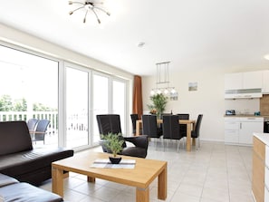 Ein modernes Wohnzimmer mit Sofa, Esstisch mit Stühlen, einer Küchenzeile und großen Fenstern.