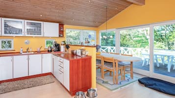 A modern kitchen with wooden cabinets, a dining area with a table and chairs, and large windows offering a view of the outdoors.