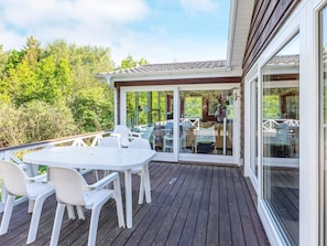 A covered outdoor deck with a table and chairs, a glass door leading to a room, and a view of greenery.