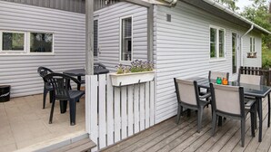 Eine überdachte Terrass mit Tisch und Stühlen, einem weißen Zaun und einem Fenster mit Blumenkasten.