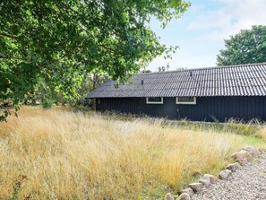 Un bâtiment rustique en bois, surmonté d’un toit en tôle ondulée, entouré d’herbes hautes et d’arbres.