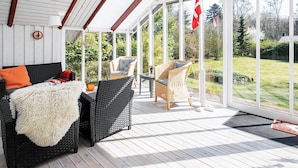 A covered patio with wicker furniture, a white fluffy throw, and a view of a garden and trees.