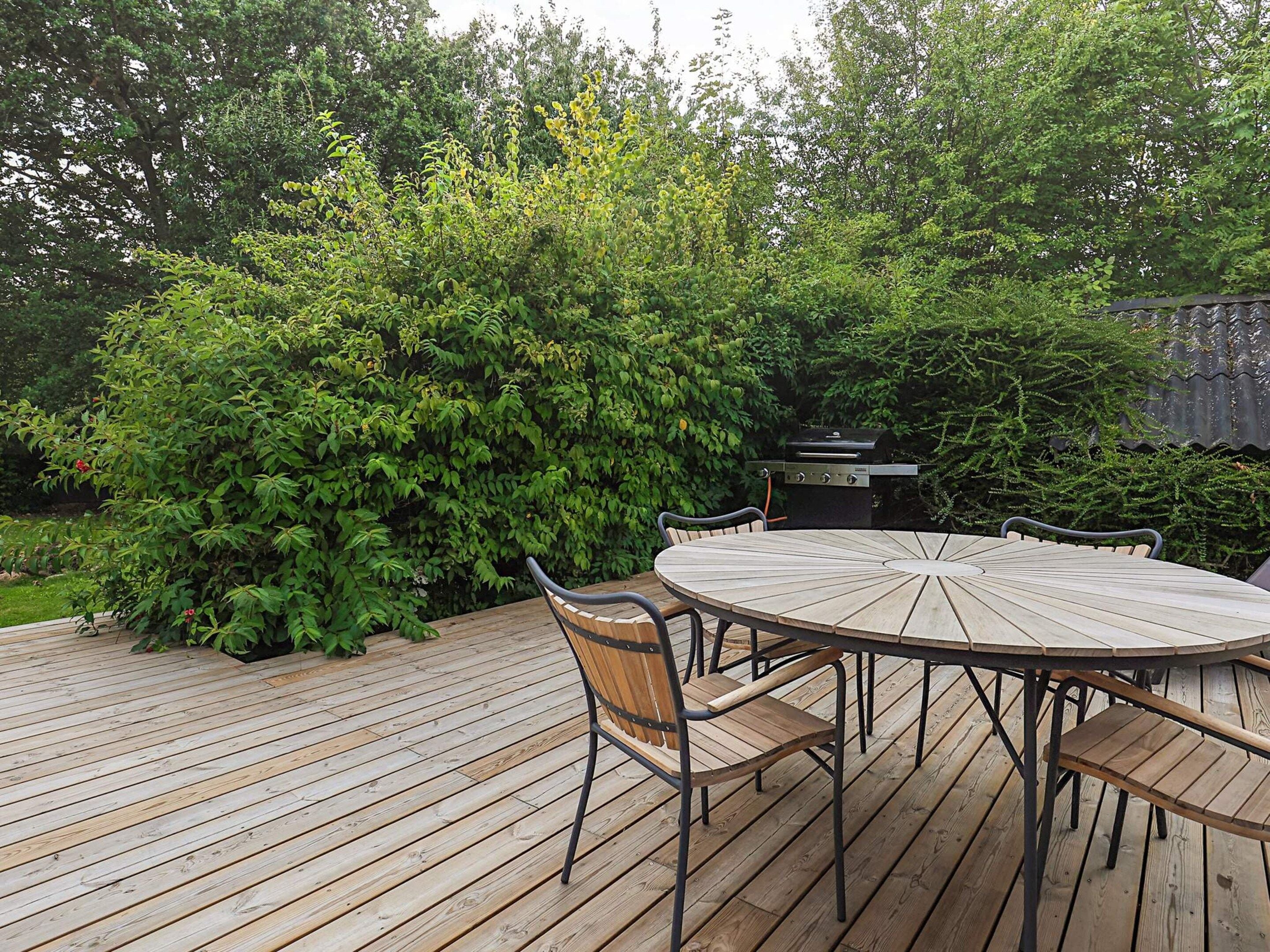 A round wooden deck with a table and chairs, surrounded by dense green foliage and a barbecue grill.