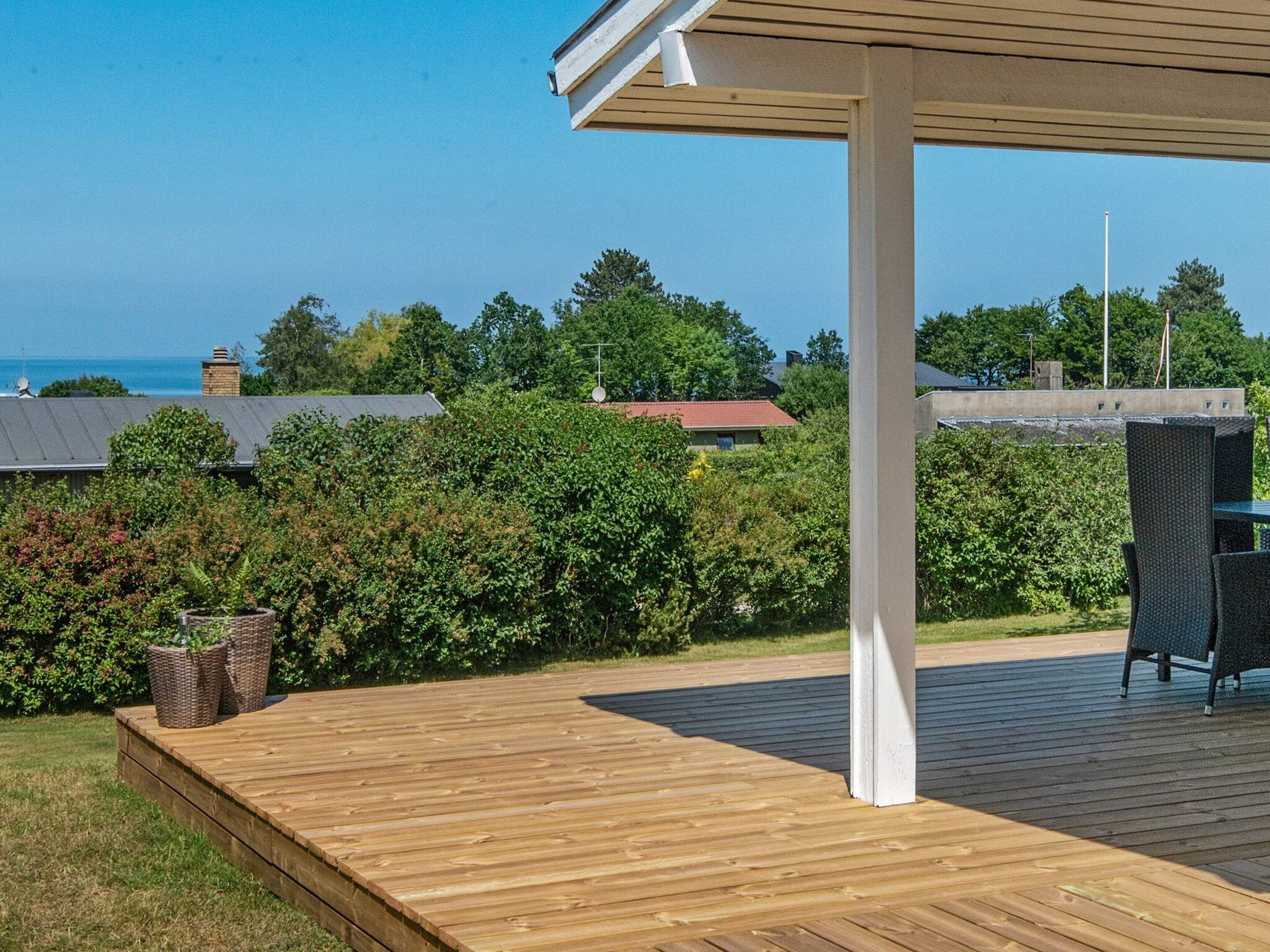 A wooden deck with a covered seating area, surrounded by greenery and a view of houses and the sea.