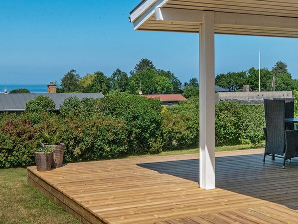 A wooden deck with a covered seating area, surrounded by greenery and a view of houses and the sea.