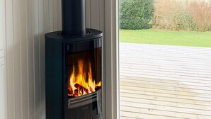 A black wood-burning stove with visible flames, positioned next to a window with a view of a wooden deck and greenery outside. - 6 Person Holiday Home in Allingabro-by Traum (Allingåbro)