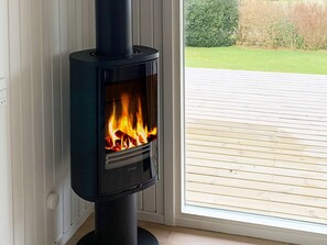 A wood-burning stove with a glass door, positioned next to a window with a view of a wooden deck and greenery outside.