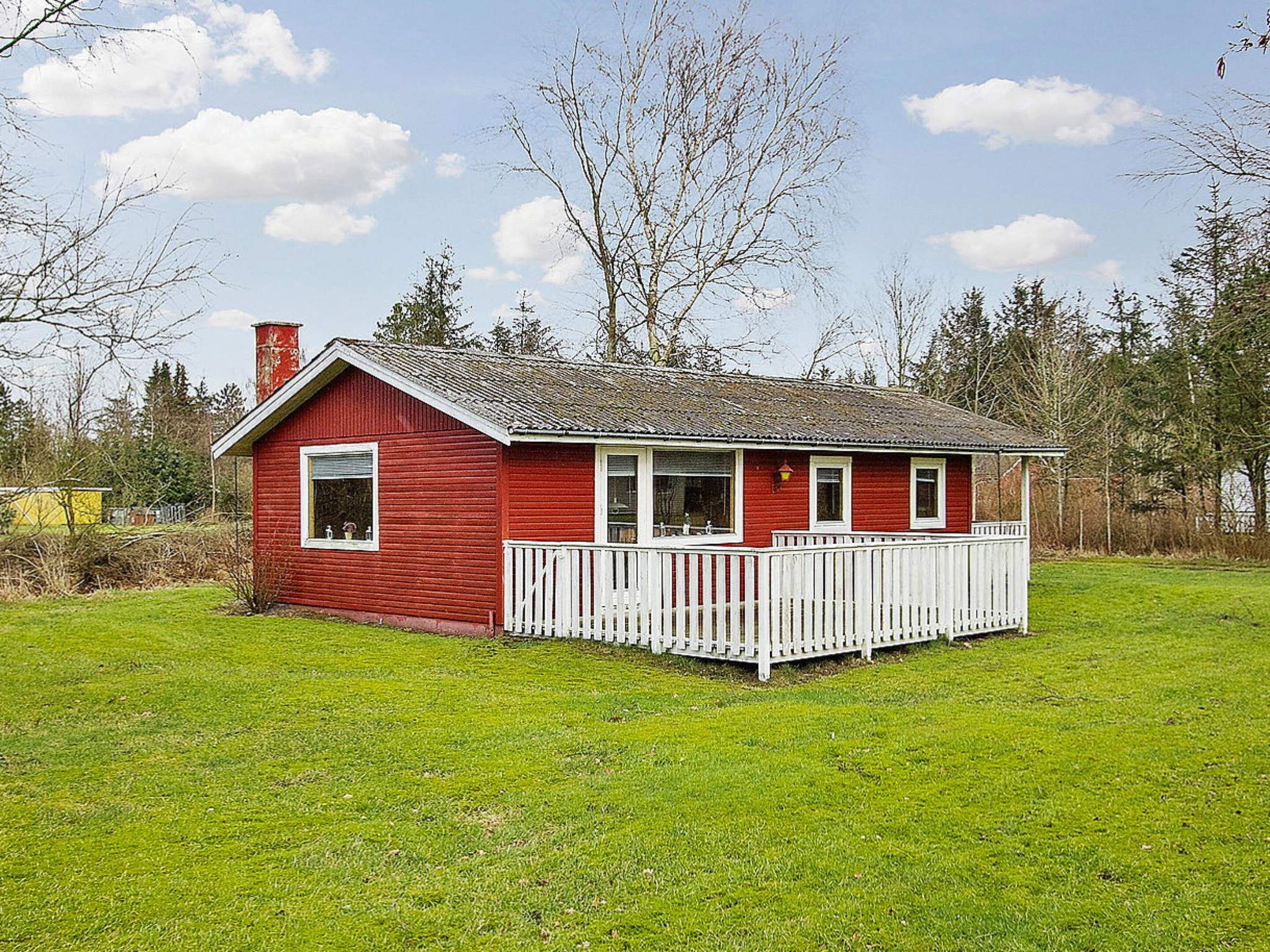 A red wooden house with a white porch and a chimney, surrounded by a grassy field and trees.