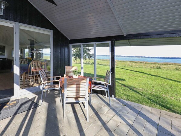 An outdoor patio with a table and chairs, a view of a grassy field and water body, and a building with a dark exterior and white trim. - 4 Person Holiday Home in Hojslev (Hojslev)