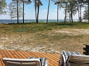 Une vue sur la plage avec une terrasse en bois, deux chaises longues et une zone herbeuse qui mène à la mer.