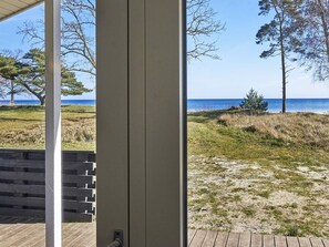 Vista desde una habitación con terraza de madera que da a una playa con vegetación escasa y cielo despejado.