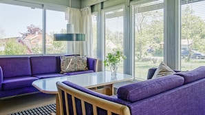 A room with a purple sofa, a round table, and large windows with blinds. - 8 Person Holiday Home in Allinge (Allinge)