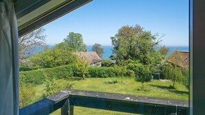 A view from a balcony with a wooden railing, overlooking a grassy area with trees and a distant view of the sea. - 8 Person Holiday Home in Allinge (Allinge)