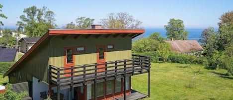 A two-story wooden house with a balcony, surrounded by a grassy yard and trees.
