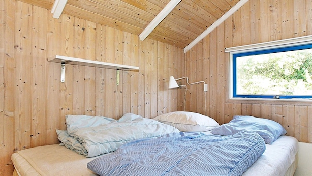 A room with a wooden interior, a bed with two pillows, a bedside table, and a window with a view of greenery.