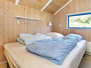 A room with a wooden interior, a bed with two pillows, a bedside table, and a window with a view of greenery.