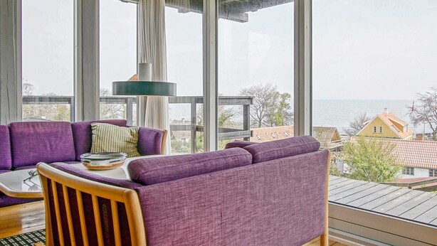 Una habitación con ventanas amplias, un sofá morado, una silla de madera y una mesa con un bol y vasos.