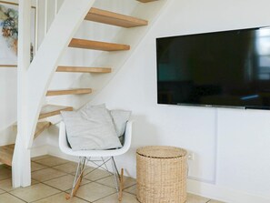 Un salon moderne avec un escalier, un téléviseur à écran plat, un fauteuil blanc et un panier en osier.