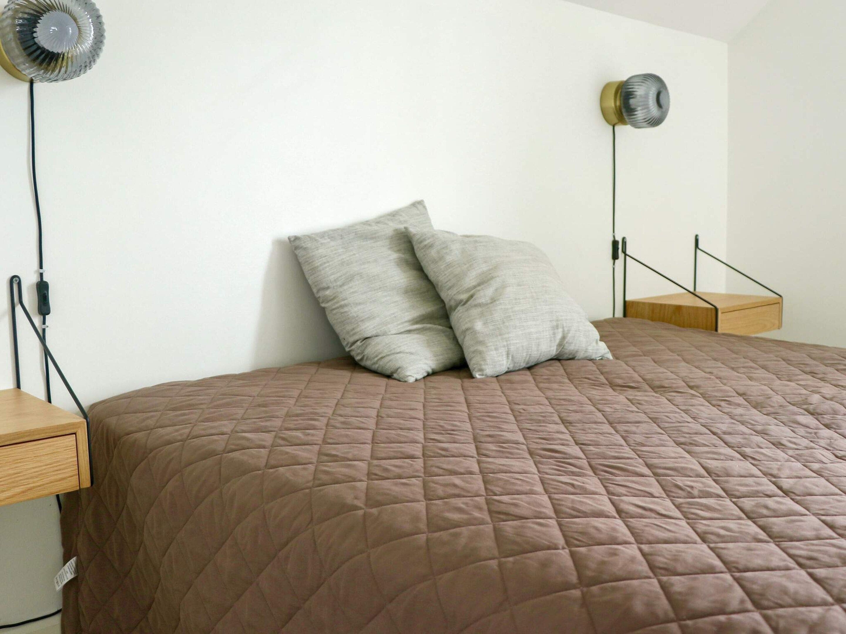 Una cama con cubierta acolchada marrón, dos almohadas, una mesita de noche, un ventilador de pared y una lámpara de noche pequeña.