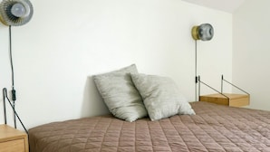 Una cama con cubierta acolchada marrón, dos almohadas, una mesita de noche, un ventilador de pared y una lámpara de noche pequeña.