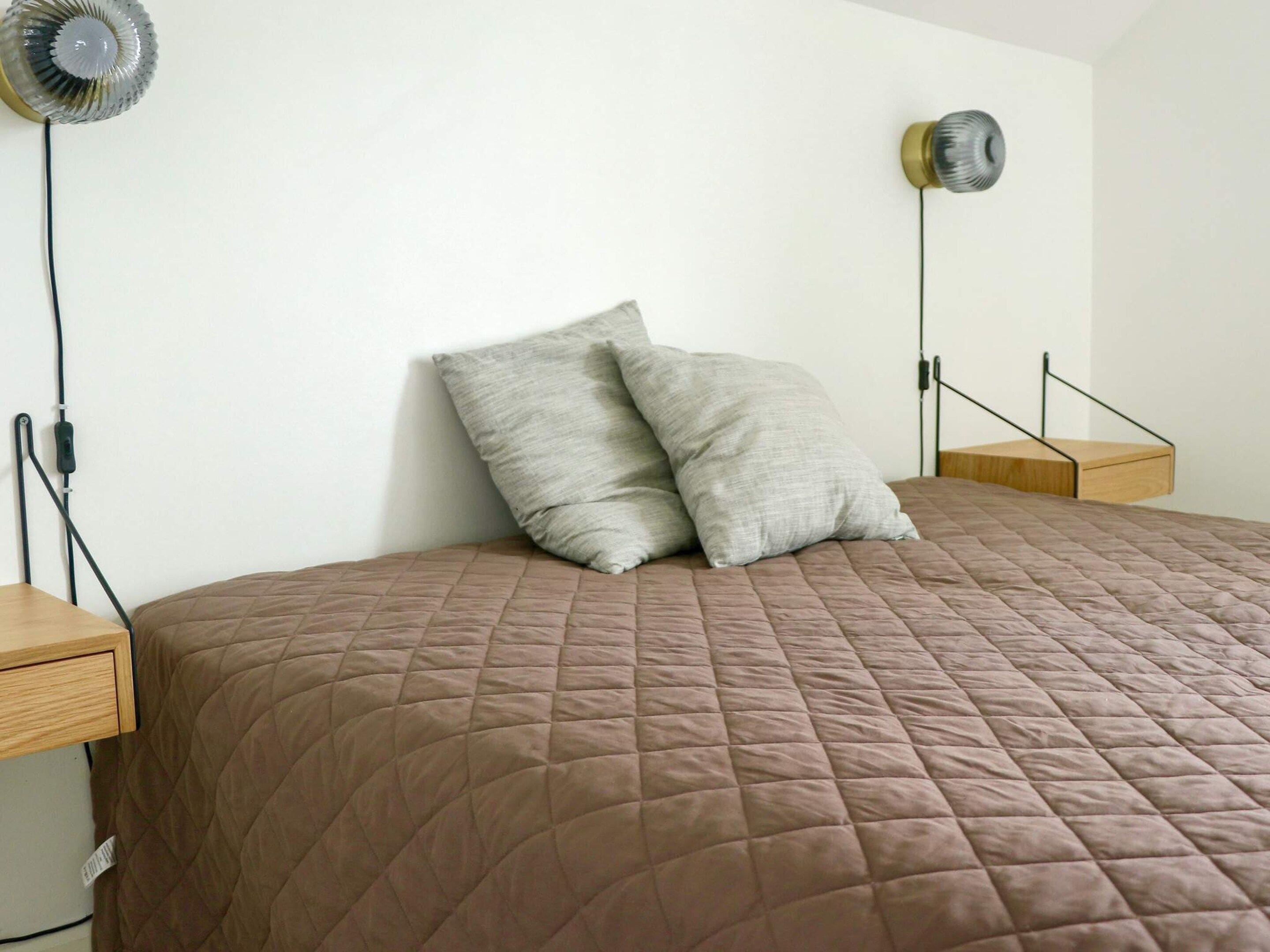 A bed with a brown quilted bedspread, two pillows, a bedside table, and a wall-mounted lamp.