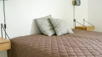 A bed with a brown quilted bedspread, two pillows, a bedside table, and a wall-mounted lamp.