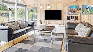 A modern living room with a wooden ceiling, a large flat-screen TV, two sofas, a coffee table, and a view through large windows.