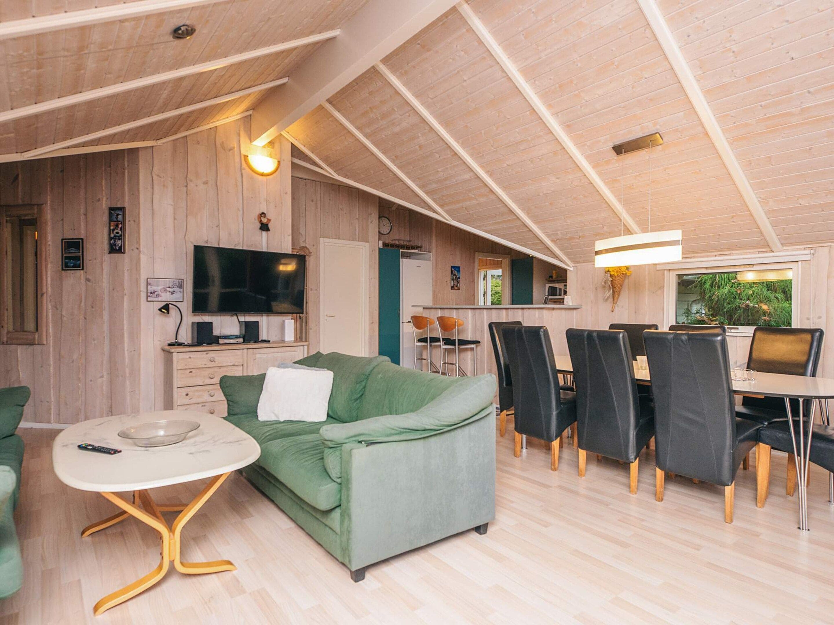 A cozy living area with a green sofa, a dining table with chairs, a TV, and wooden walls.