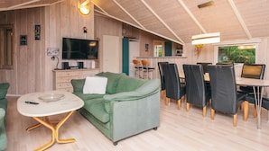 A cozy living area with a green sofa, a dining table with chairs, a TV, and wooden walls. - Pleasing Holiday Home in Ebeltoft near Sea (Ebeltoft)