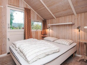 Un dormitorio con paneles de madera, una cama, dos ventanas con cortinas, una mesita de noche y una lámpara de pared.