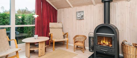 Um quarto acolhedor com lareira a lenha, cadeiras de vime, uma mesa redonda e vista através de grandes janelas.