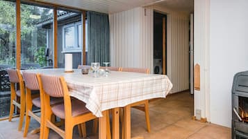 A dining area with a table set for four, wooden chairs, a window with curtains, and a view of greenery outside.