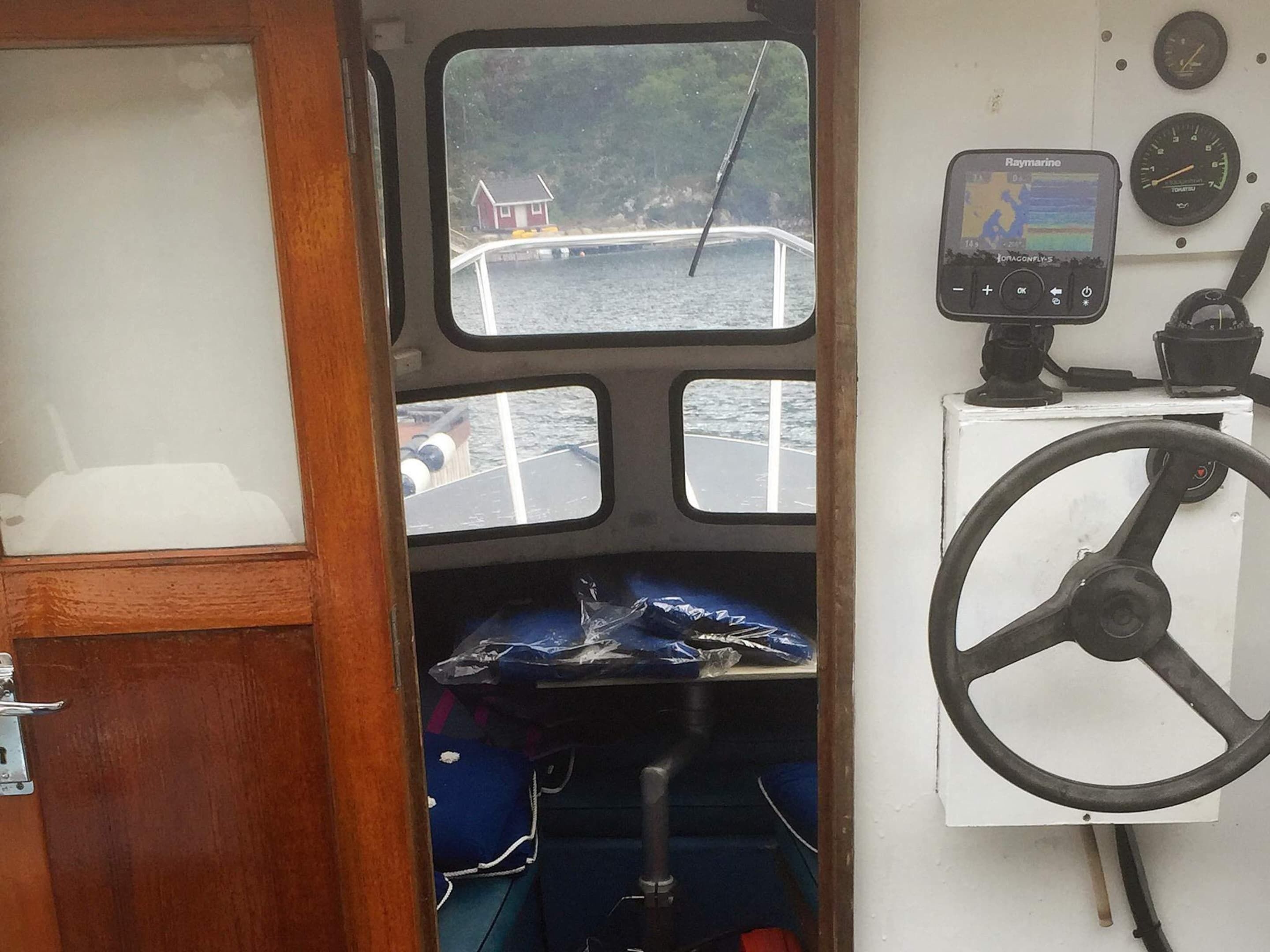 A boat's interior with a steering wheel, a Raymarine navigation device, and a view of the water through a window.